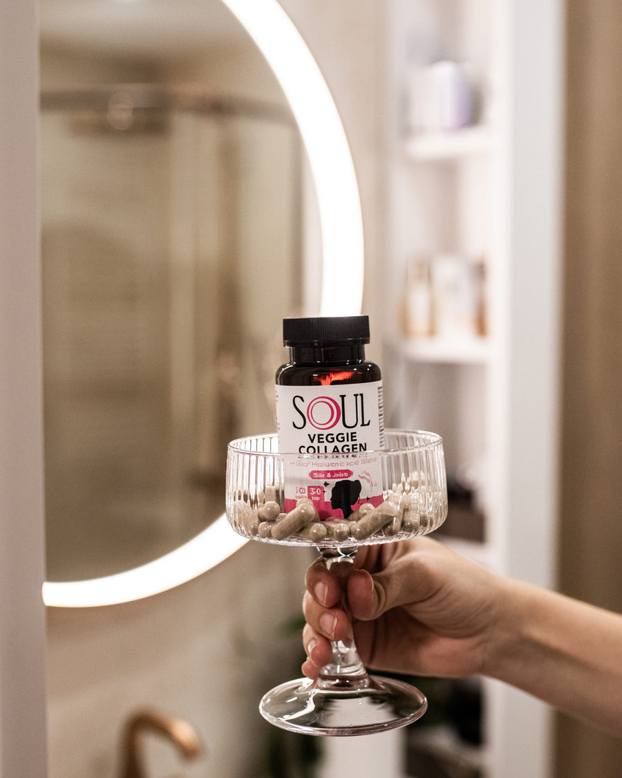 A hand holds a glass bowl of capsules and SOUL Veggie Collagen in front of a lit mirror